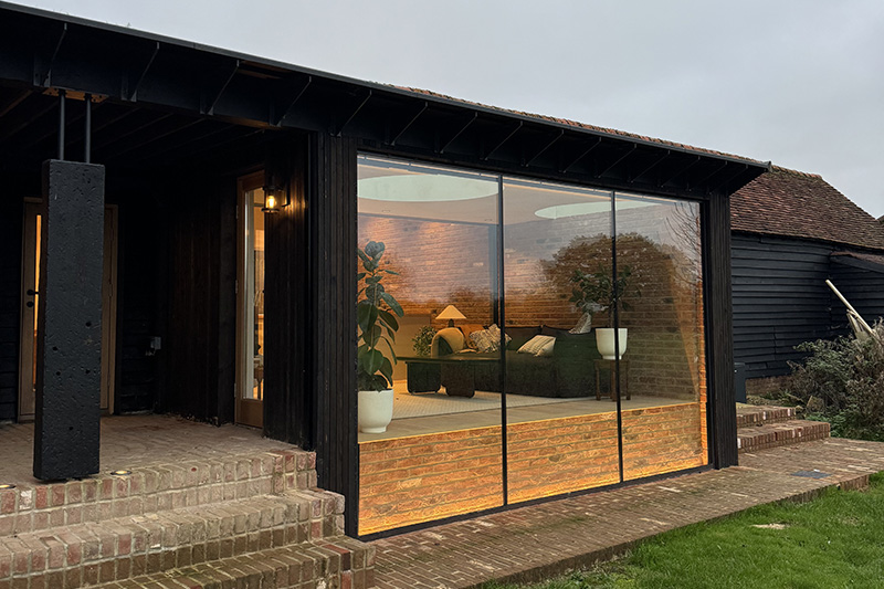 Black timber-clad barn elevation with full-height glazing onto a warmly lit living room at dusk